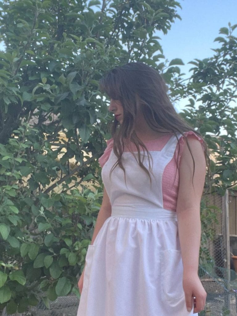 A girl with an apron on standing in the backyard of a suburban homestead.