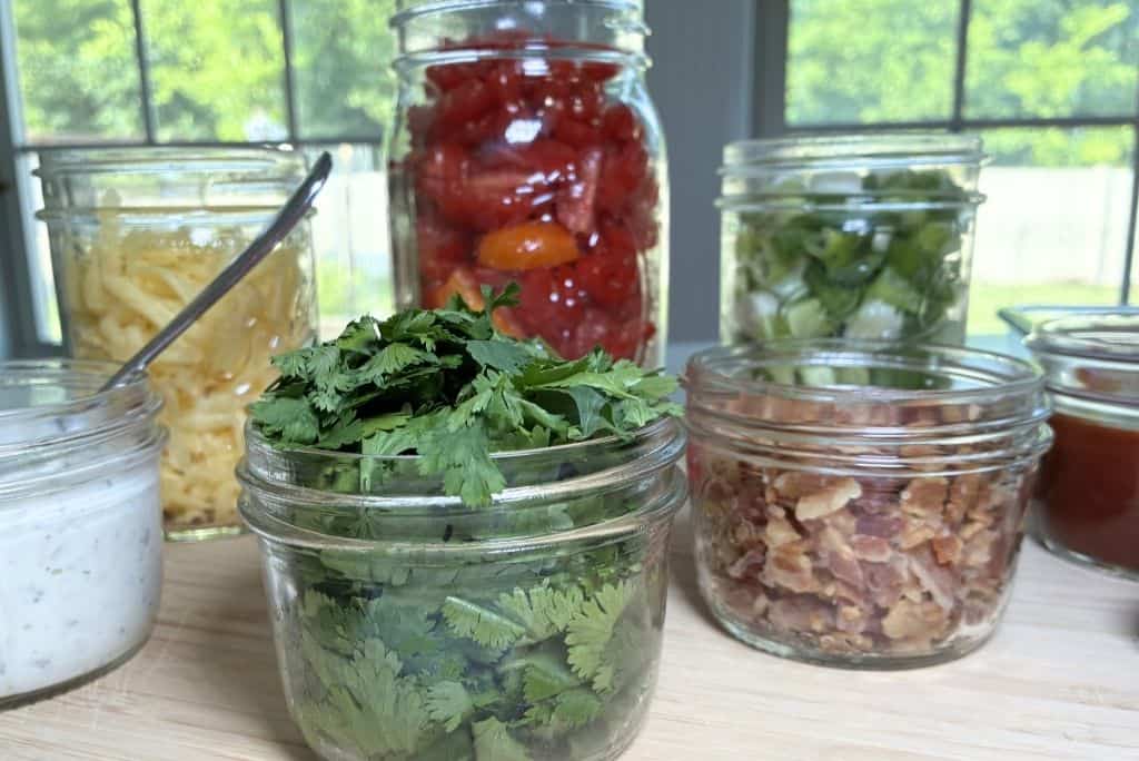 Small, medium, and large Mason jars with ranch dressing, barbeque sauce, bacon pieces, cilantro leaves, sliced green onions, chopped tomatoes, and shredded cheese.