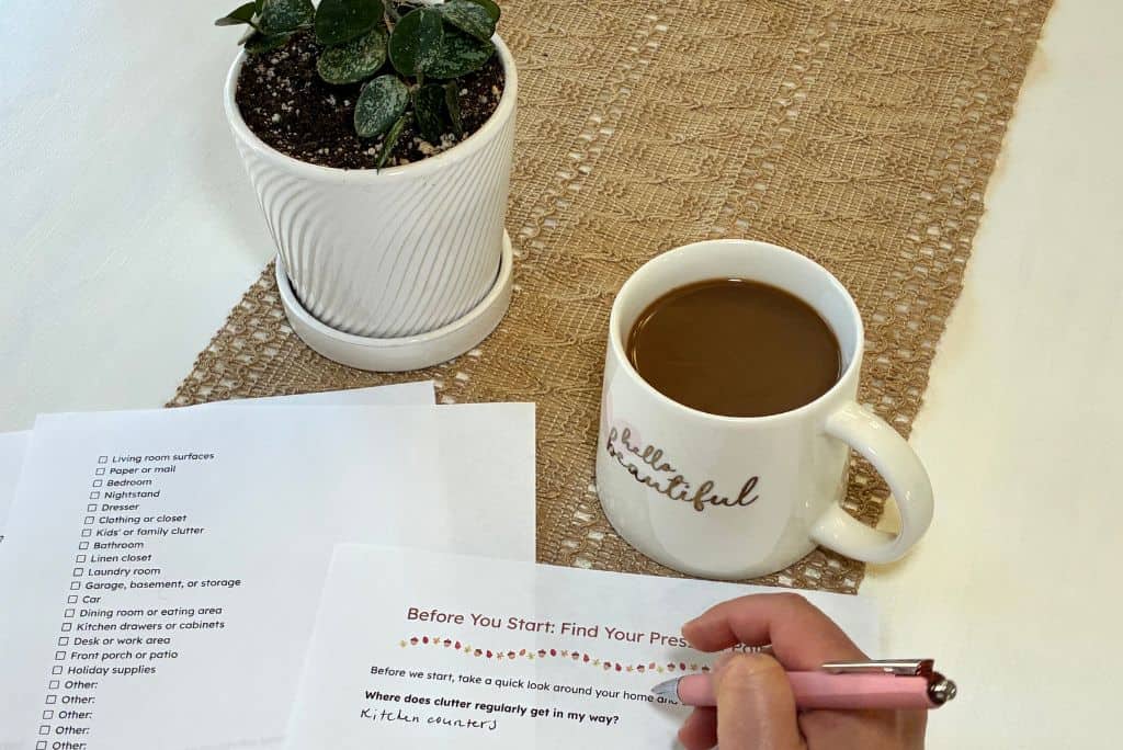 My hand filling out planning worksheets next to a potted plant with my coffee mug on my kitchen table.